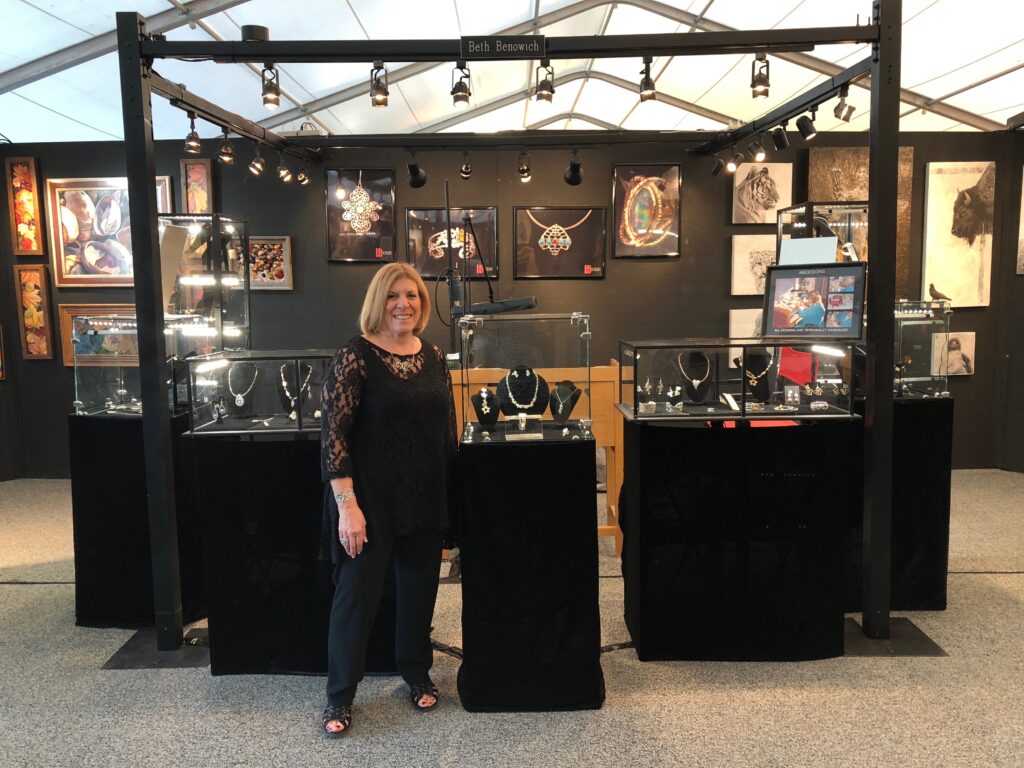 Beth Benowich of BBDesigns in a professional art show booth with illuminated glass display cases of handcrafted necklaces and earrings. | Scottsdale Luxury Lifestyle Magazine | life +style magazine | L+S